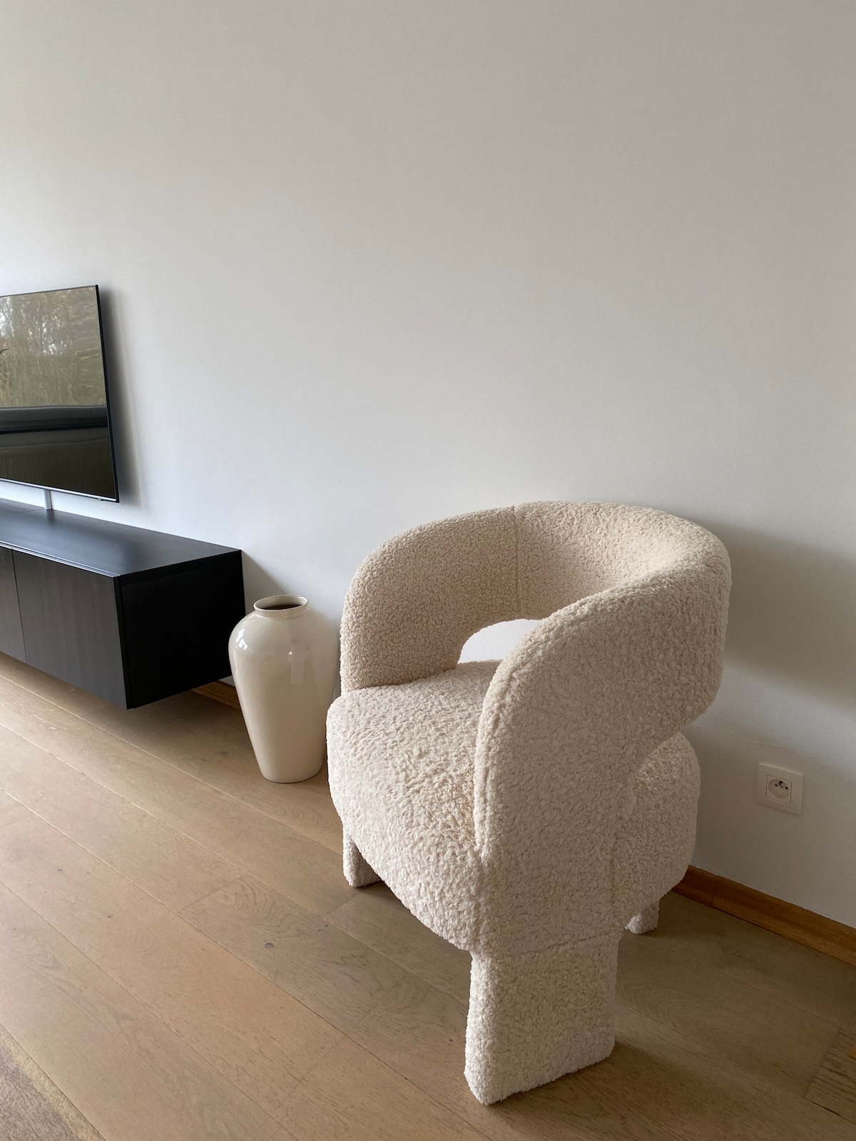 A textured chair with a rounded backrest and soft upholstery is positioned next to a low black media console. A sleek white vase stands beside the chair against a clean, light-colored wall, complementing the warm wooden flooring.