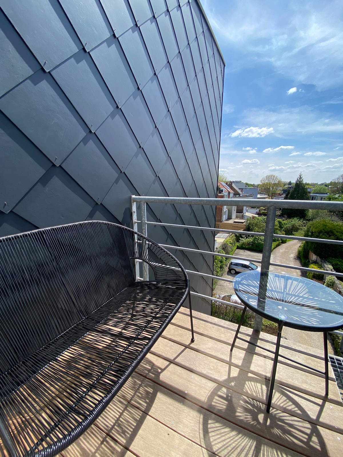 A small balcony is depicted, featuring a black, woven seating area paired with a matching round table. Natural light from the clear sky enhances the space, while a view of the surrounding greenery is visible below.
