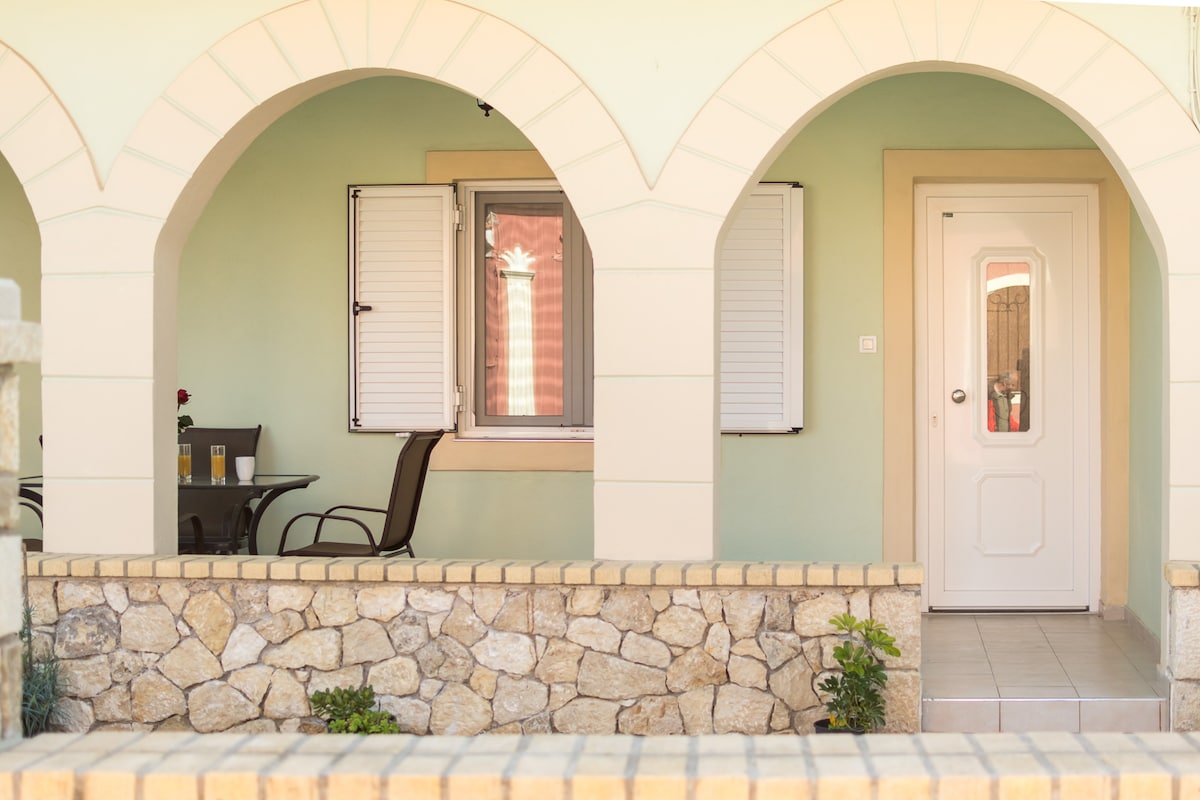An outdoor covered area features an arched design with a stone wall perimeter. A small table is set for two, accompanied by two chairs. Light-colored walls complement the shutters on the window, which adds to the inviting ambiance. A white door provides access to the interior.