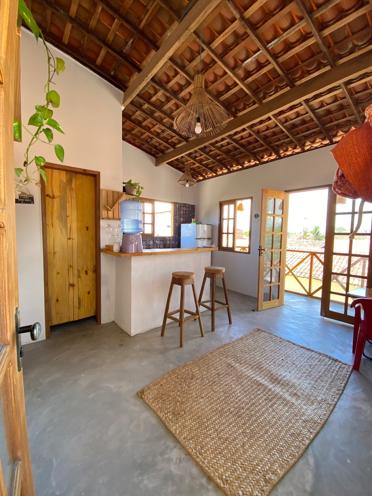 The kitchen area features a unique wooden ceiling with decorative light fixtures. A small bar with two wooden stools is positioned near a water dispenser. A textured rug adds warmth to the concrete floor, while large windows provide natural light and views of the surroundings.