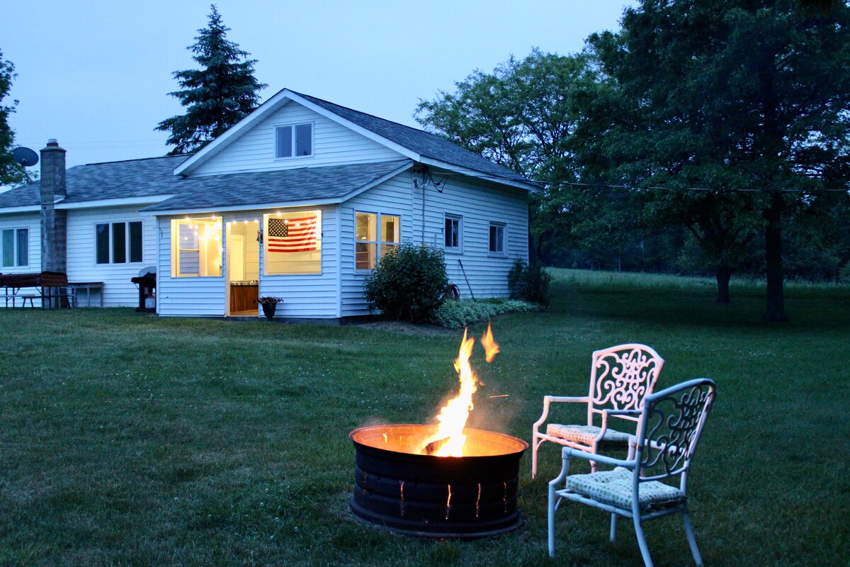 An inviting outdoor space features a fire pit surrounded by grassy land, with a single white chair positioned nearby. The charming cottage is visible in the background, illuminated by soft lighting at dusk, showcasing a welcoming atmosphere.