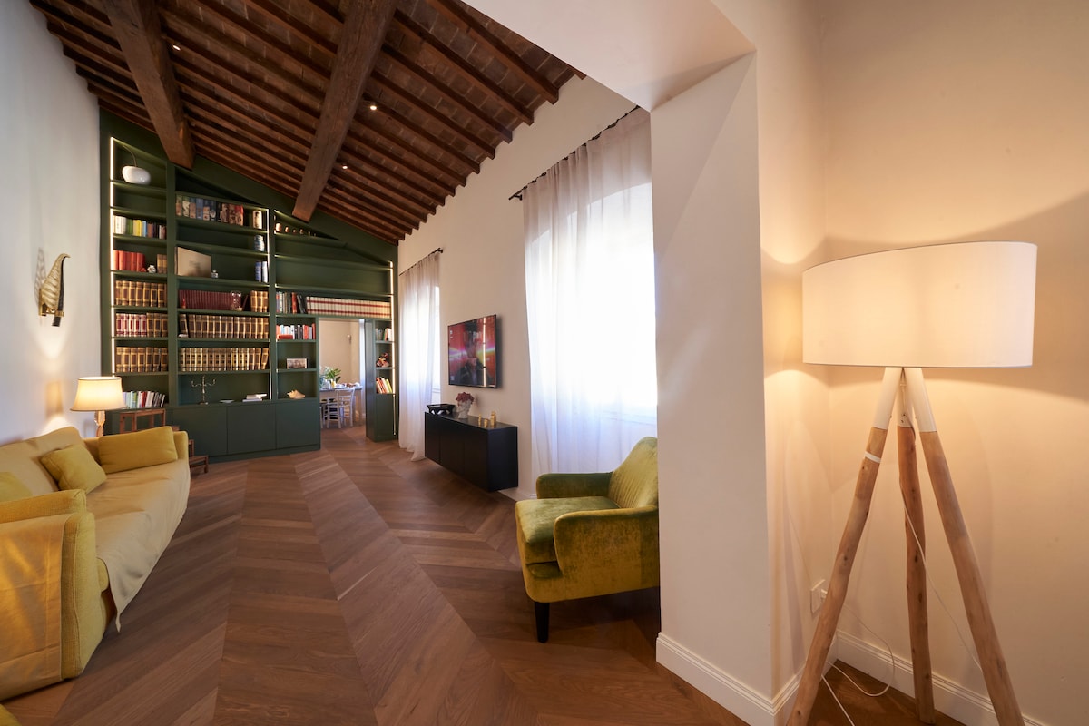 The spacious living area showcases exposed wooden beams and warm parquet flooring. A soft green sofa and a bright yellow chair complement the decor. Natural light enters through large windows, illuminating a modern entertainment center with a flat-screen TV and decorative shelving filled with books.