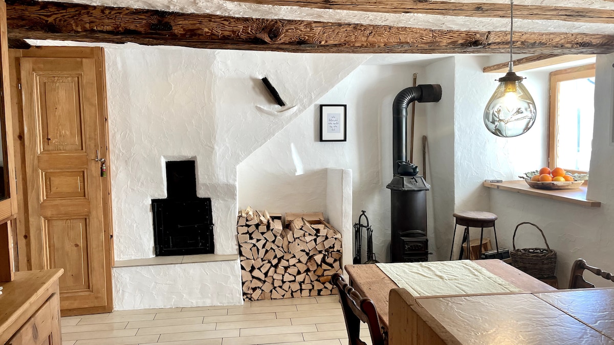 A well-appointed kitchen area features rustic wooden beams and warm-toned cabinetry. A wood-burning stove is positioned beside a neatly stacked pile of firewood. A dining table is set, with natural light illuminating the space from the nearby window.
