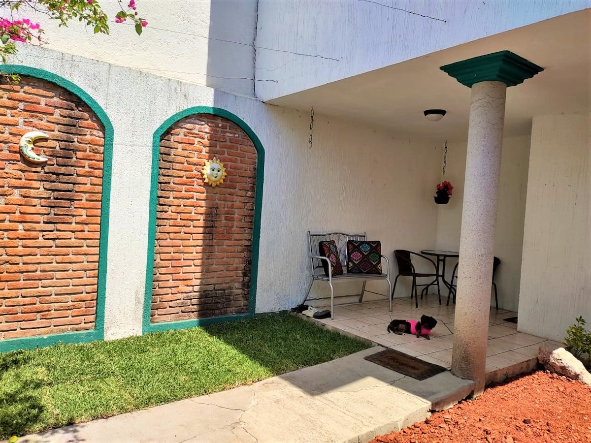 A charming outdoor space is displayed, featuring a small seating area with a table and chairs under a shaded porch. Two brick walls adorned with decorative sun and moon motifs create a rustic feel, while green grass and vibrant flowers add a touch of nature.