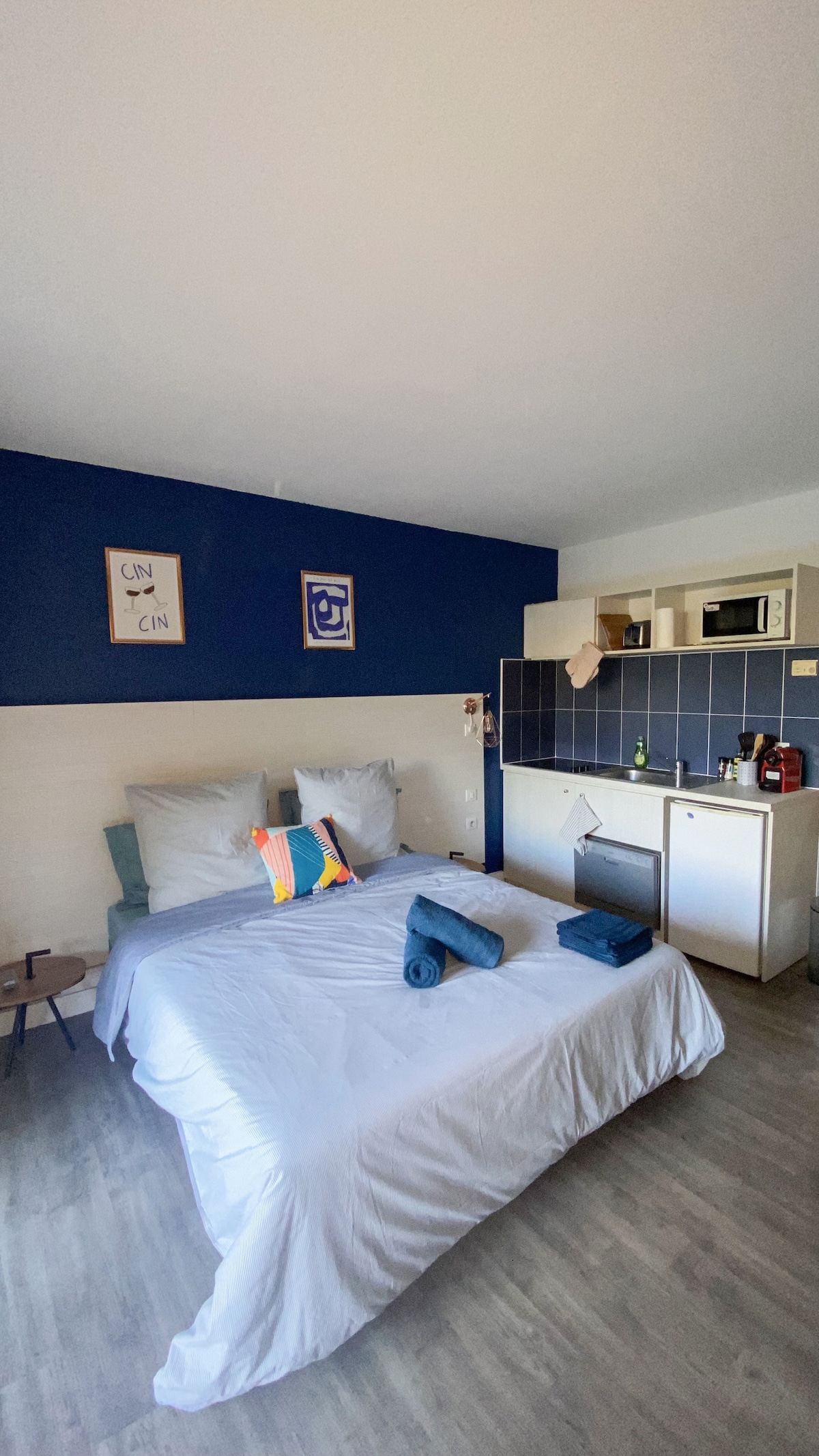 A studio with a cozy sleeping area is showcased, featuring a spacious bed with crisp linens and decorative pillows. The kitchenette, equipped with a refrigerator and cooking amenities, is visible in the background. Two framed artworks adorn the blue accent wall, complementing the neutral tones of the decor.