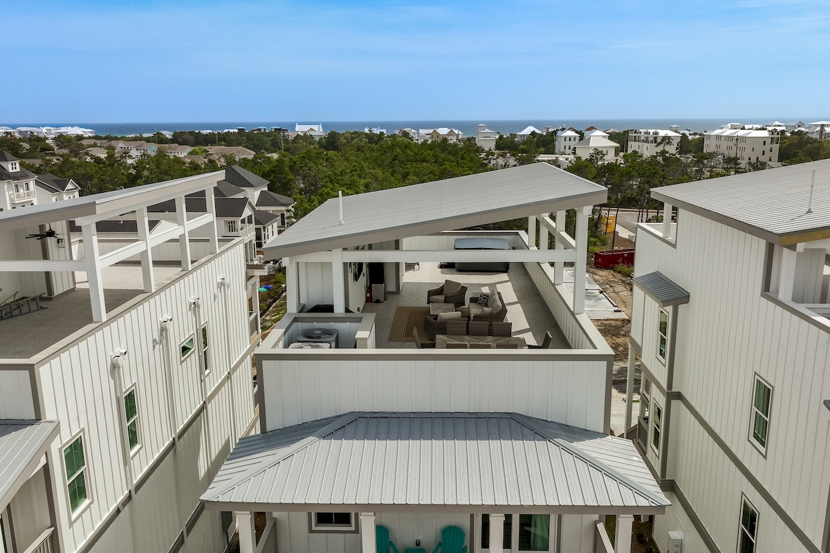 An aerial view captures a coastal home with a modern rooftop deck that offers sweeping Gulf views. The deck features a comfortable seating area and is accessible from the upper levels of the building, surrounded by a neighboring landscape of homes.