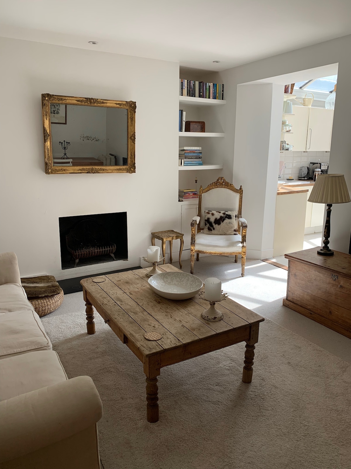 A cozy living area features a soft sofa and a rustic wooden coffee table, accompanied by a decorative bowl. A golden-framed mirror hangs above a black fireplace. A vintage armchair adds character, while books are neatly arranged on nearby shelves, enhancing the inviting ambiance.
