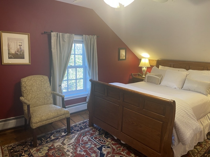 A cozy guest room features a spacious bed with white linens, complemented by a decorative lamp on a nightstand. A comfortable chair sits beside the bed, while natural light streams in through a window dressed with sheer curtains, reflecting off the rich red walls.
