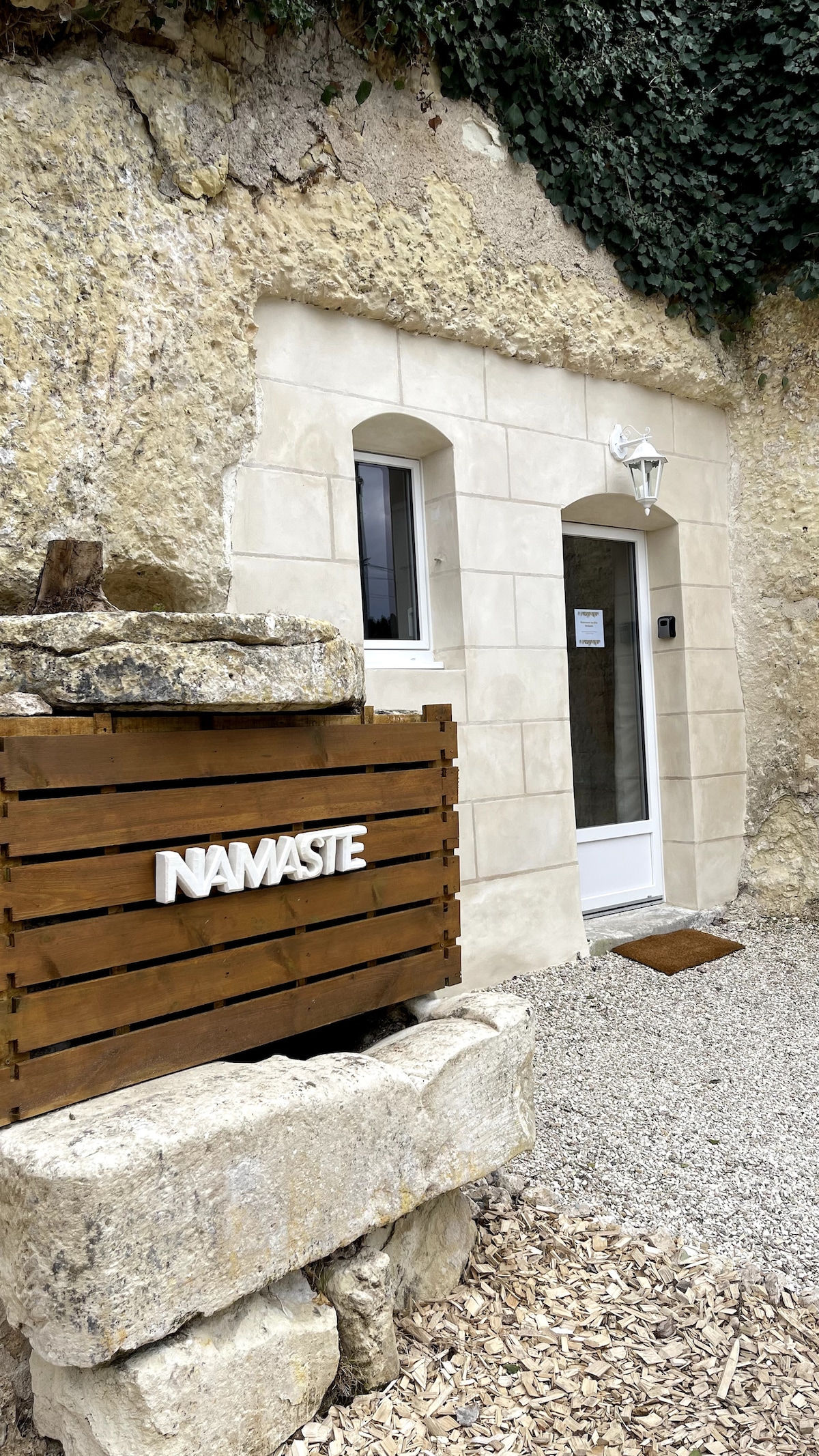 The entrance of the troglodyte studio is framed by natural stone walls, with an arched window above the door. A wooden sign reading 'NAMASTE' is mounted on a rustic wooden panel. The path is lined with gravel, enhancing the serene outdoor setting.