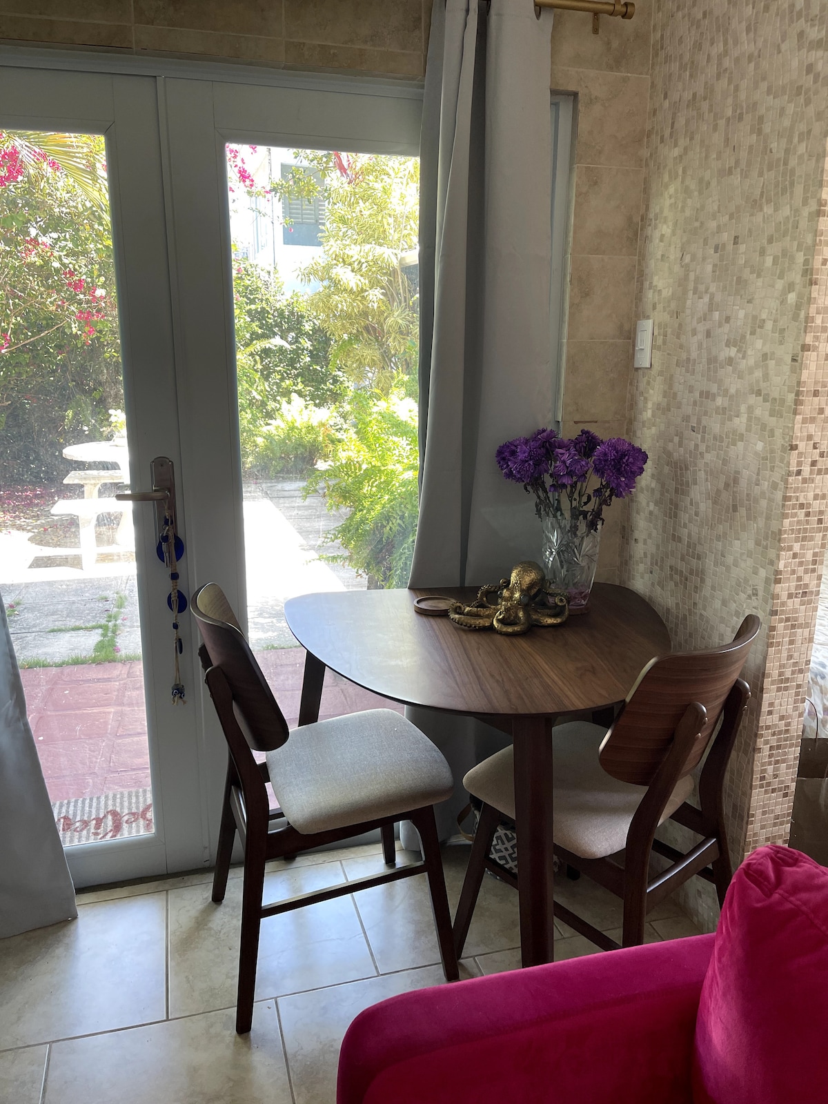 A small dining table is set against a glass door that opens to a garden. Two wooden chairs accompany the table, which is adorned with a vase of purple flowers. Natural light filters through the curtains, illuminating the space.