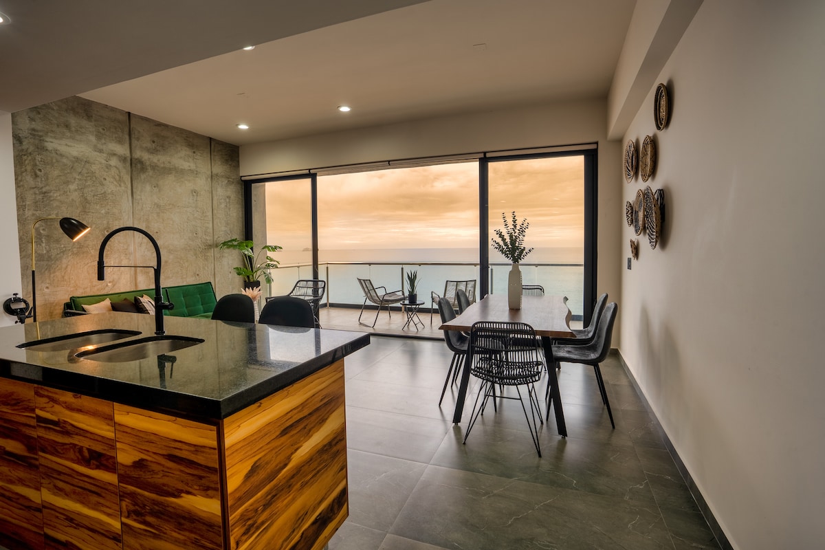 An open-concept living area features a kitchen with a black countertop and wooden cabinets. Large glass doors reveal a stunning sunset view over the ocean, while a dining table and chairs are positioned nearby, complemented by modern decor on the wall.
