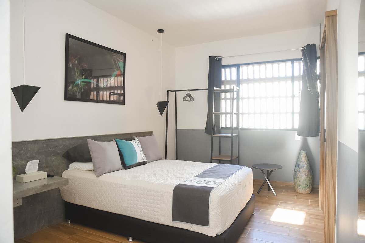 A comfortable bedroom features a spacious bed with neutral-toned bedding and decorative pillows. A sleek hanging clothing rack stands nearby, while a small round table is positioned beside the bed. Natural light filters through the window, which is framed by dark curtains.
