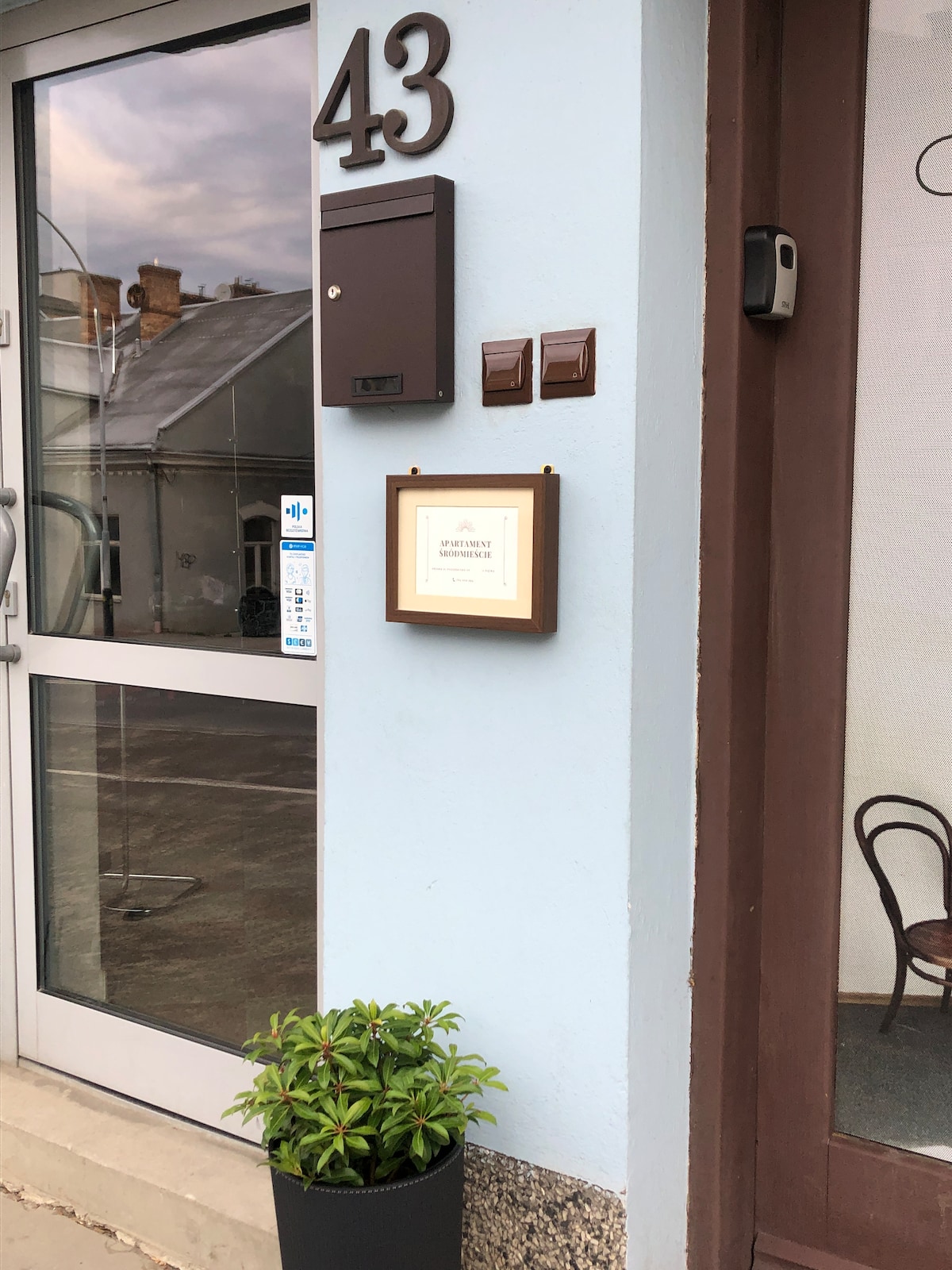 The entrance displays a light blue wall with a large number '43' prominently featured. A modern mailbox is mounted beside a framed sign. A simple plant in a black pot rests on the ground, adding a touch of greenery to the welcoming entryway.