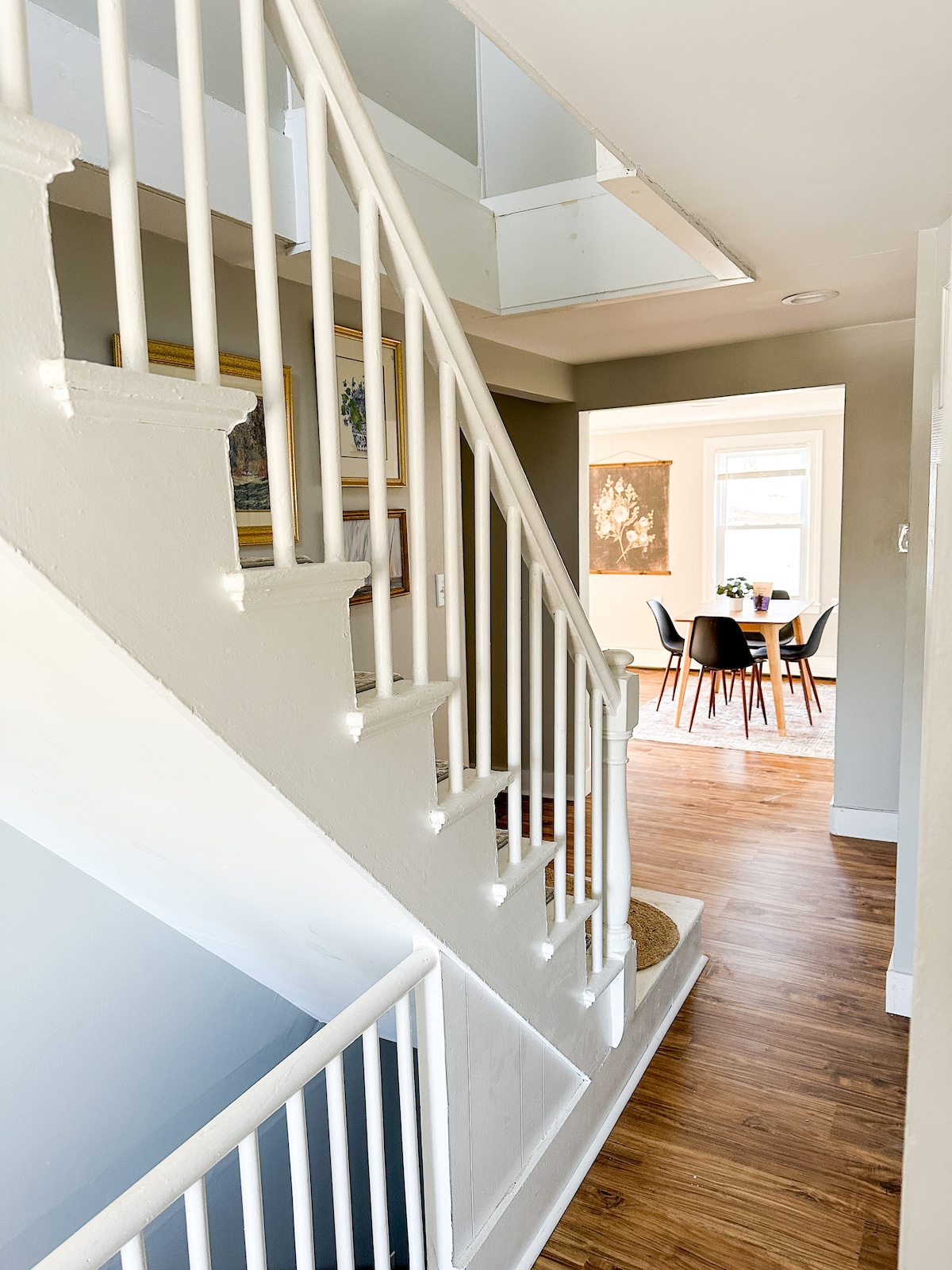 A light-filled hallway features a white staircase leading to the upper level. Light wood flooring extends through the space, which includes a glimpse of a dining area with a round table and chairs in the background.