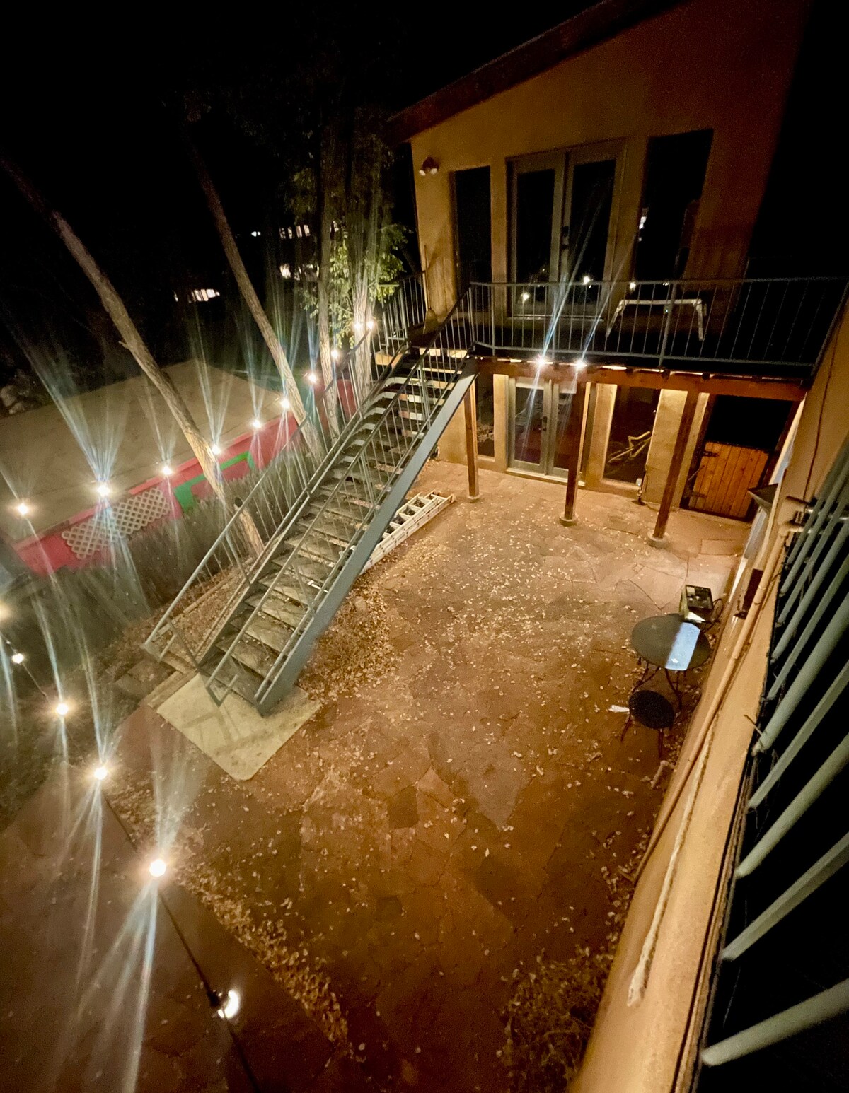 An outdoor patio area is illuminated by soft lighting, enhancing the inviting atmosphere. A staircase leads to an upper level, while the patio features a table and chairs amidst fallen leaves, surrounded by trees creating a serene setting.