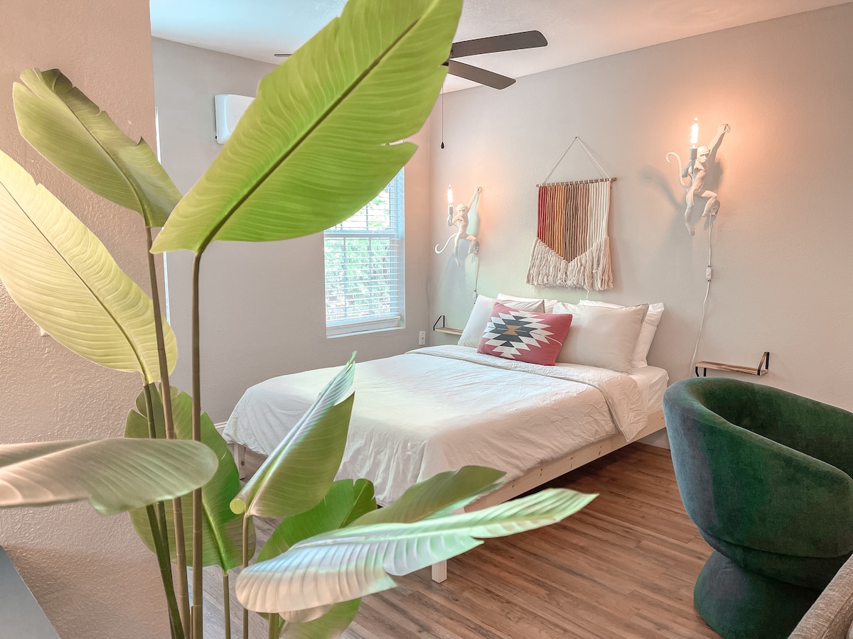 A spacious sleeping area features a comfortable bed adorned with crisp white linens and decorative pillows. Natural light enters through a window, while stylish wall sconces provide soft illumination. A green chair and large leafy plant add a touch of freshness to the inviting space.