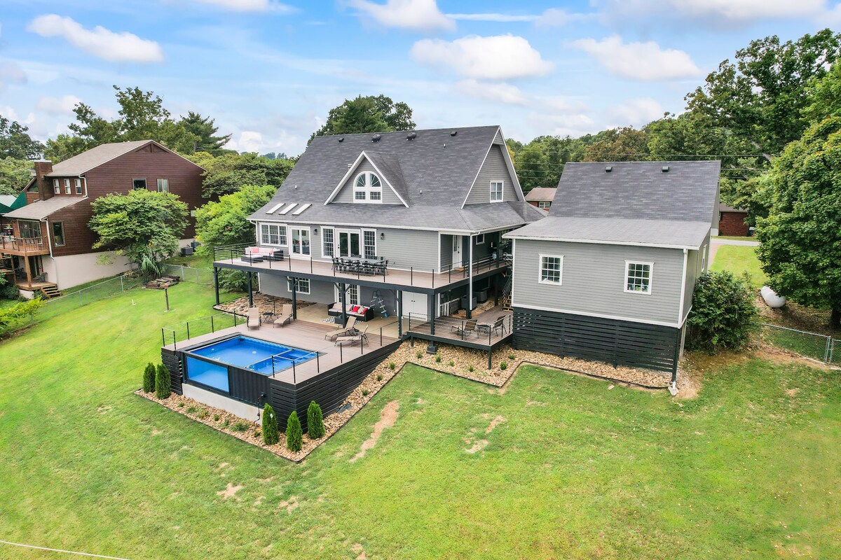 An aerial view reveals a spacious two-story home with a modern design, surrounded by lush greenery. A heated or chilled pool is positioned in the backyard, accompanied by a deck and patio area, showcasing furniture for relaxation and gatherings.