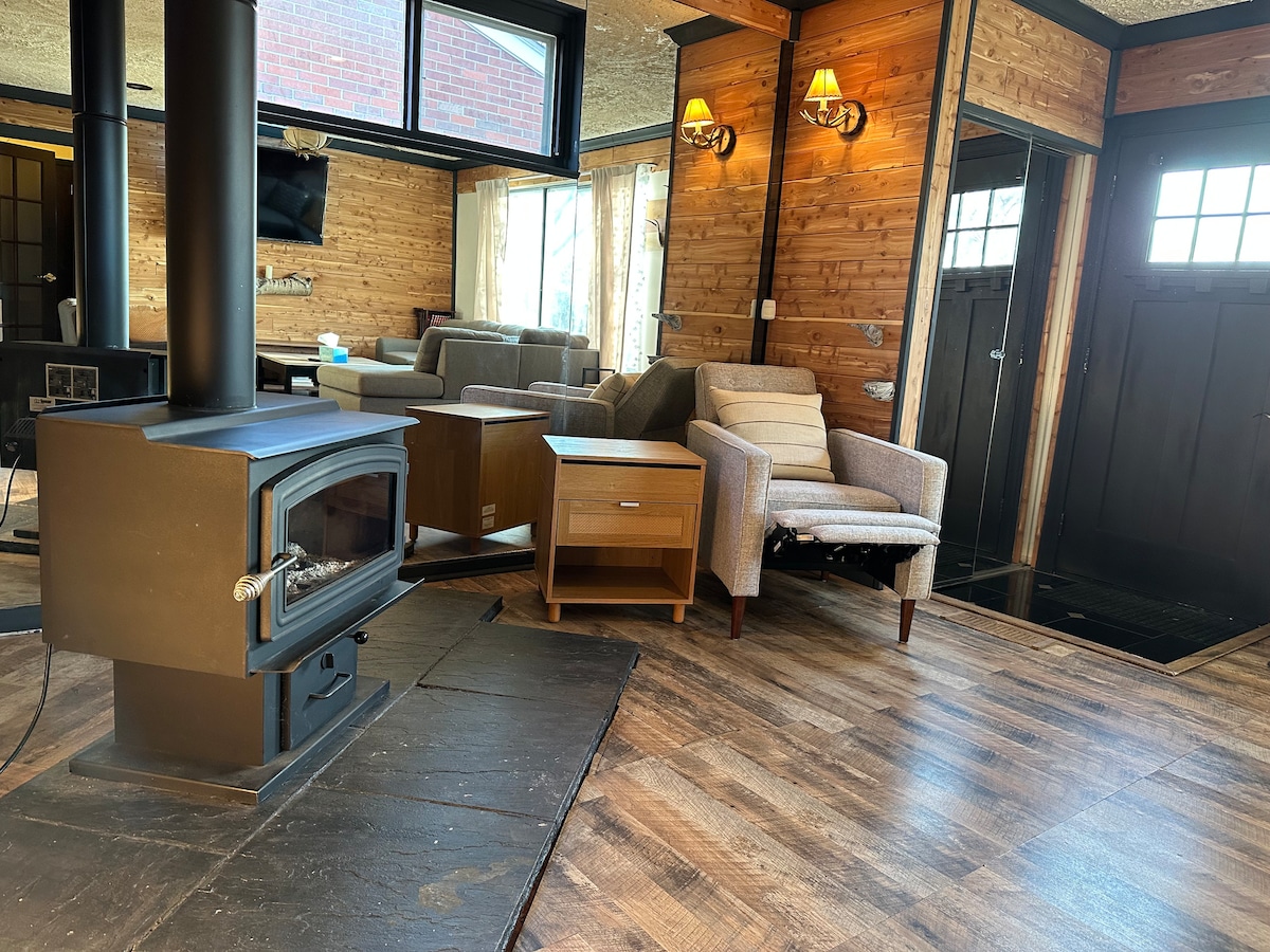 A cozy living space is highlighted by a wood-burning stove positioned against a backdrop of cedar walls, offering warmth. Comfortable seating is arranged around a small table, with large windows allowing natural light to fill the room, creating a serene atmosphere.