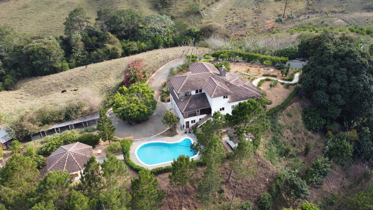 An aerial view showcases a spacious villa surrounded by lush greenery and rolling hills. A large, inviting swimming pool is set in the garden, complemented by several trees and flowering plants, offering a serene atmosphere. The layout reveals clear paths connecting various outdoor areas.