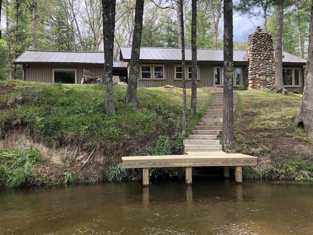 The cabin is positioned on a gentle slope, overlooking the river. A wooden dock extends into the water, providing access for fishing and relaxation. A stone chimney adds character to the rustic exterior, surrounded by lush greenery and tall trees.