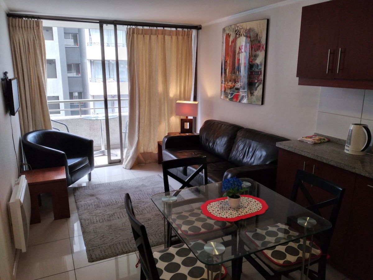 A bright living space featuring large glass doors that provide access to a balcony. A comfortable black leather sofa and an armchair are arranged around a glass dining table with patterned chairs. A lamp and decorative artwork enhance the room's design.
