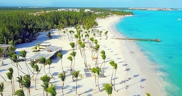 An aerial view of a sandy beach lined with palm trees, overlooking clear turquoise waters. The shoreline features people enjoying the scenery, while various structures are situated near the beach, offering shaded areas for relaxation.