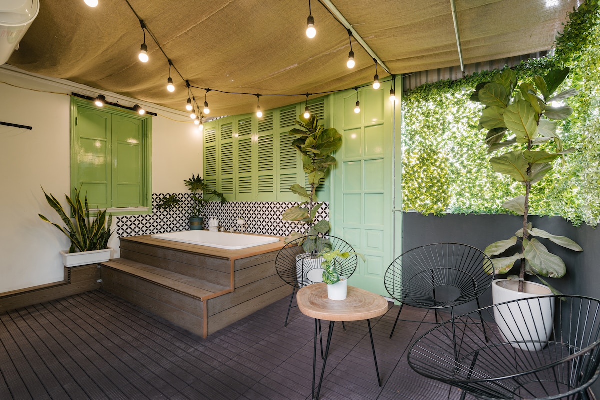 A rooftop balcony features a large, inviting hot tub surrounded by a decorative black-and-white tile wall. Comfortable seating areas are provided by black wire chairs and a small wooden table. Green plants add a touch of nature, with soft lighting enhancing the ambiance.