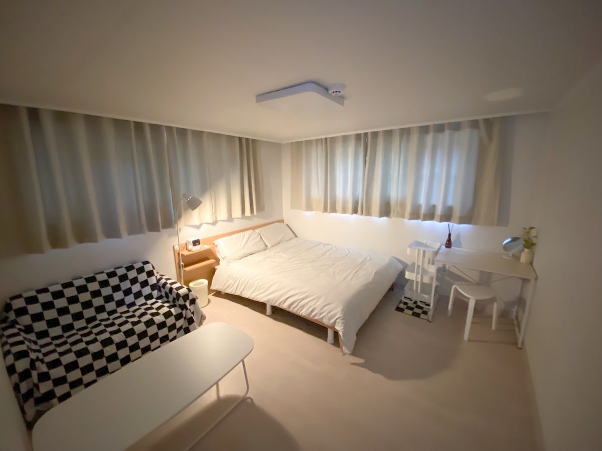A well-lit room features a spacious bed with white bedding and a checkered sofa. Natural light filters through two sets of light curtains on either side. A small table and chair are positioned next to a side table, creating a functional space for relaxation.