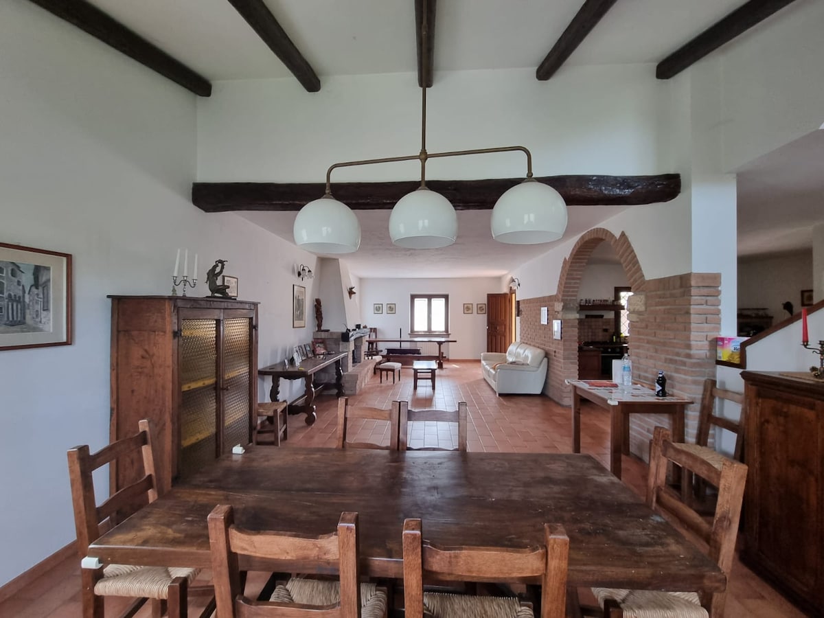 An open dining area with a large wooden table surrounded by rustic chairs is visible. Three pendant lights hang from the ceiling beams above. The room features a spacious living area and an archway leading to additional spaces, with natural light entering through a central window.