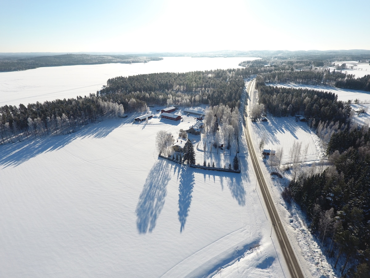 An aerial view captures a serene countryside landscape blanketed in snow, dotted with a cluster of buildings surrounded by trees. A winding road edges the property, leading toward a distant tree line and a frozen lake, creating a tranquil and expansive atmosphere.
