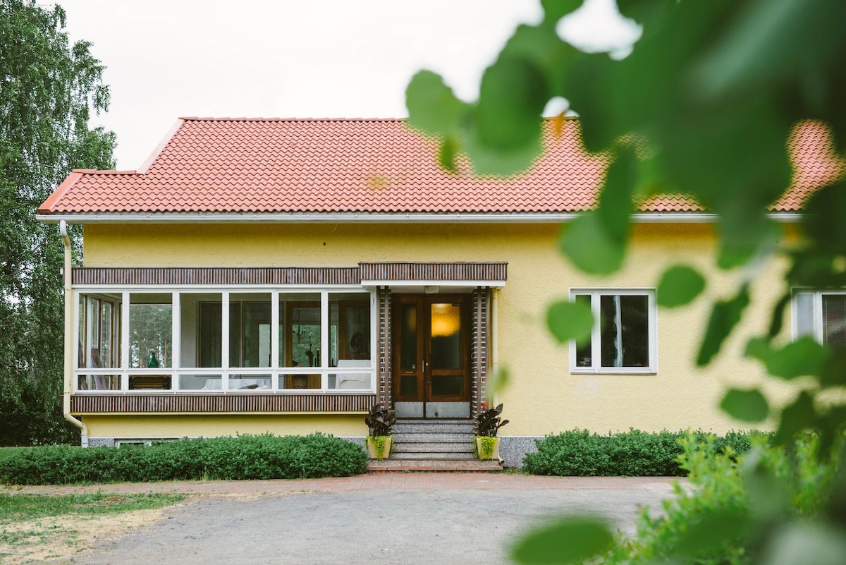 A spacious house features a bright yellow exterior with a sloped red roof. Large windows line the front, allowing ample natural light. Steps lead to the welcoming entrance, framed by lush greenery and potted plants, enhancing the home's connection to the surrounding nature.