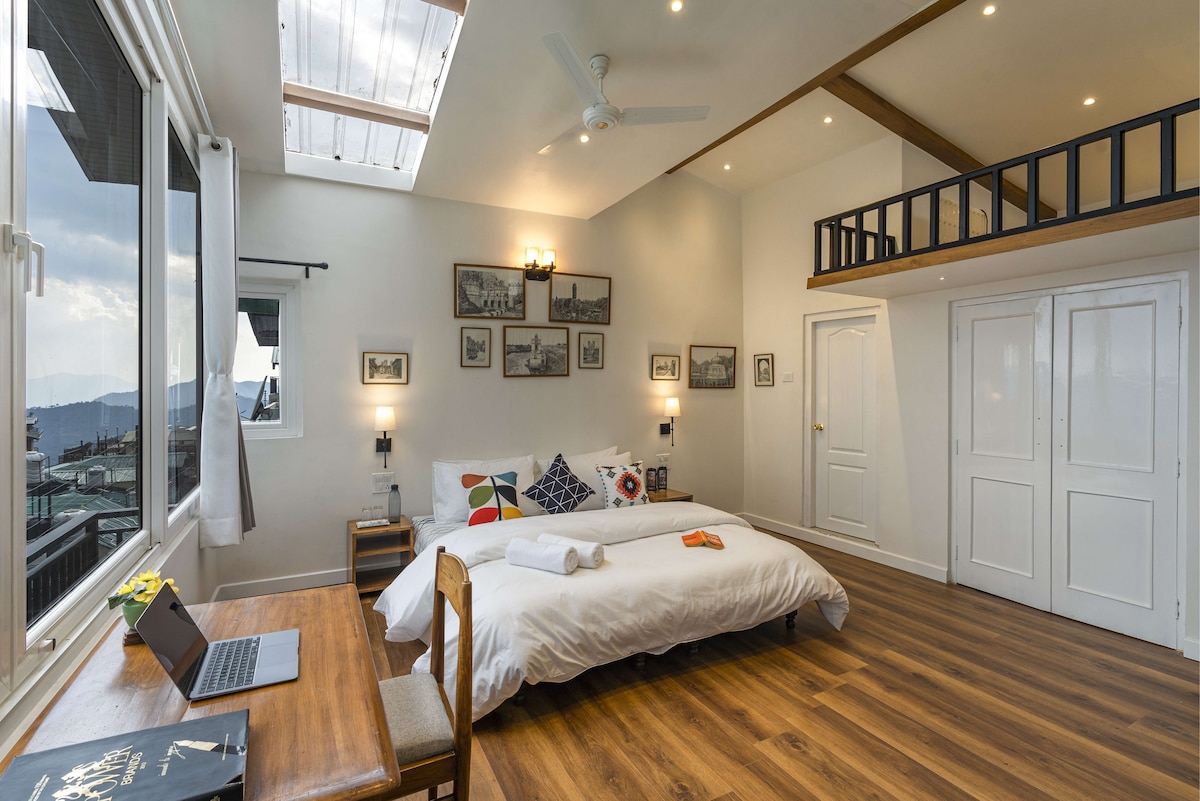 A spacious room features a large bed adorned with colorful cushions. Natural light filters through the skylight and windows. A desk and chair are positioned neatly beside the bed, while artwork and photographs decorate the walls, contributing to a homely feel.