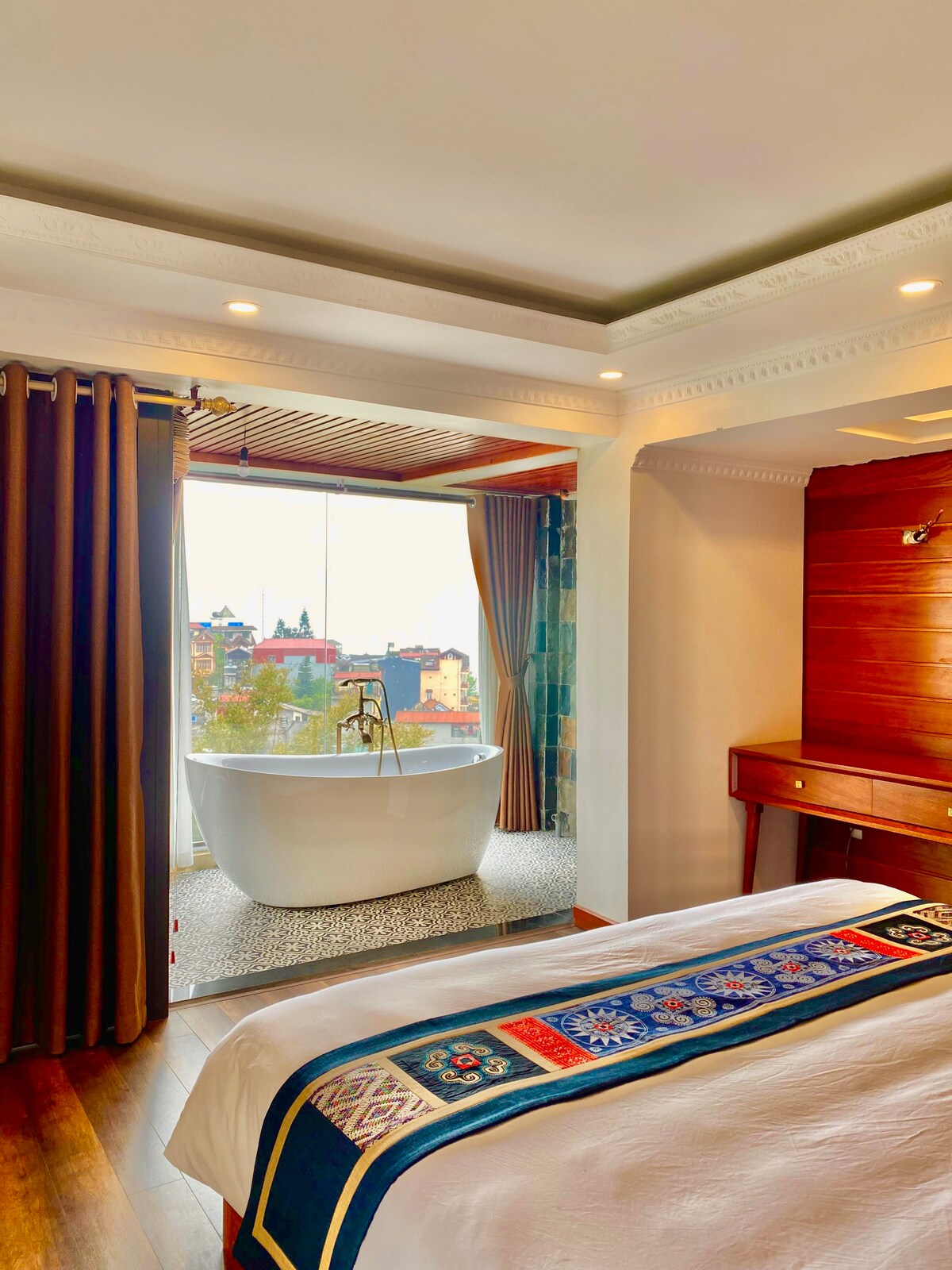 A spacious bedroom features a large white tub beside floor-to-ceiling windows, offering views of the surrounding landscape. Rich wooden accents and modern decor are present. A decorative blanket lies on the bed, complementing the room's elegant style.