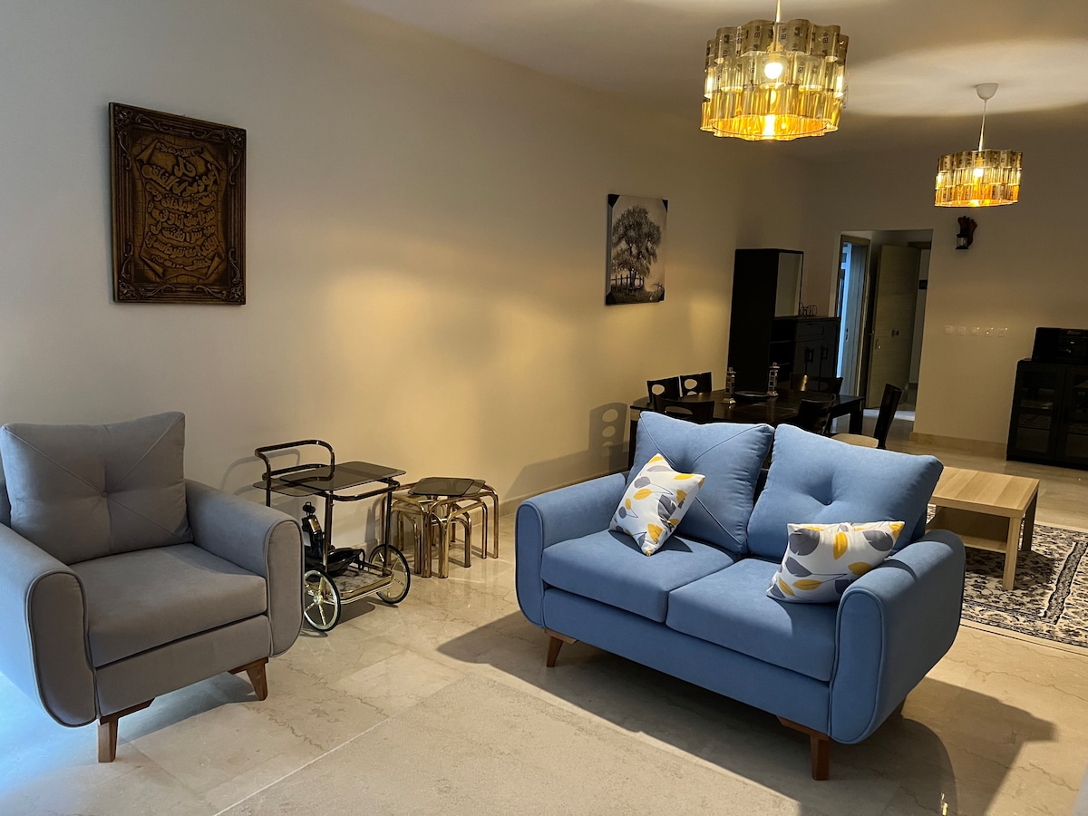A spacious living area features a light blue sofa with decorative cushions and an armchair nearby. A wooden coffee table sits in front of the sofa, while elegant light fixtures hang from the ceiling. A dining area with a dark table and chairs is visible in the background.