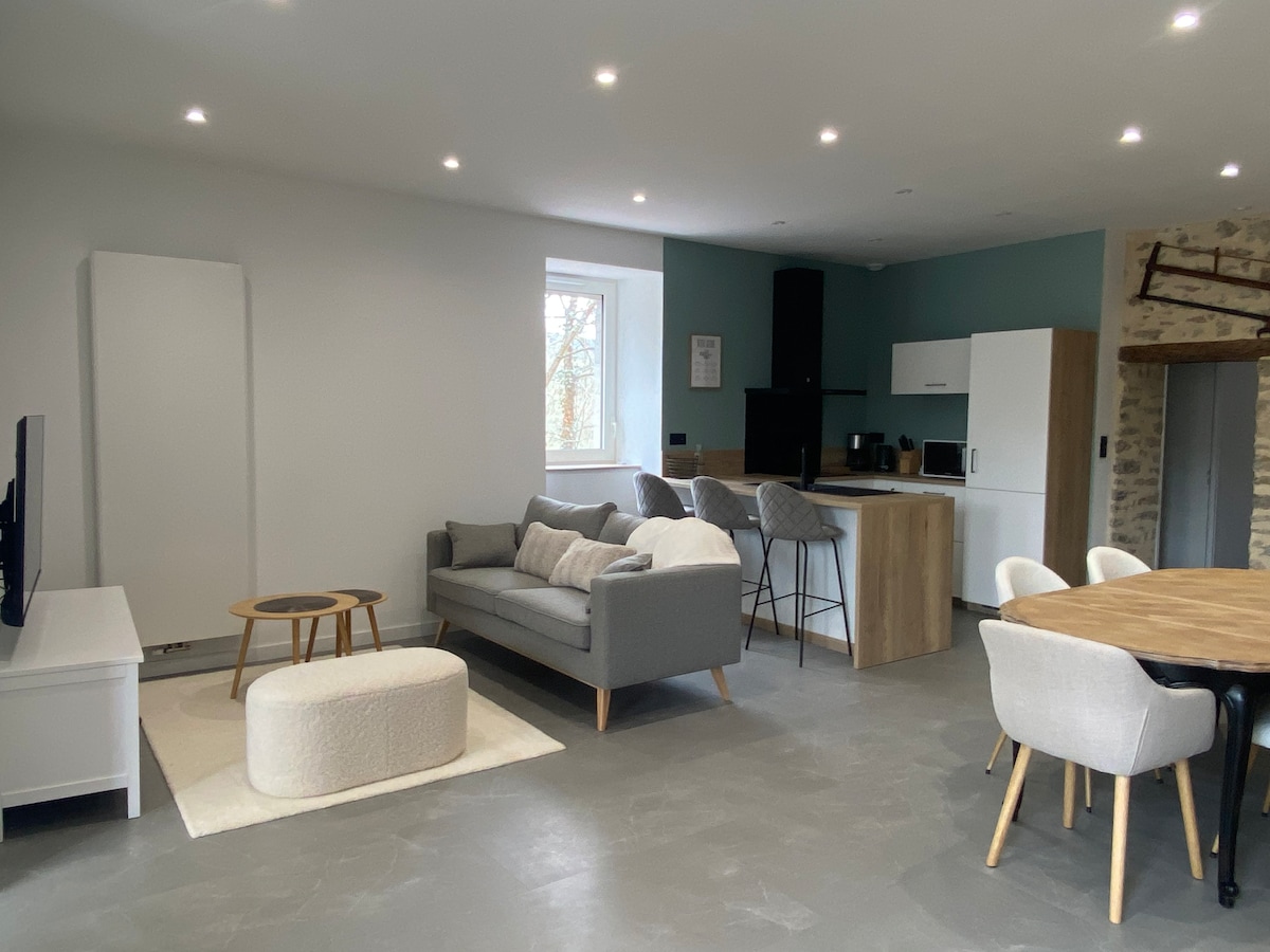 A spacious living area features a light grey sofa and round coffee table, surrounded by soft seating and a dining area. A modern kitchen with white cabinetry and black appliances is visible in the background, complemented by natural light from a nearby window.