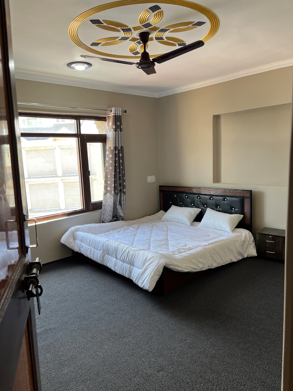 A well-lit bedroom features a spacious bed with a dark headboard. Natural light comes through a large window adorned with patterned curtains. The room includes a bedside table, and soft carpet covers the floor, contributing to the overall comfort of the space.