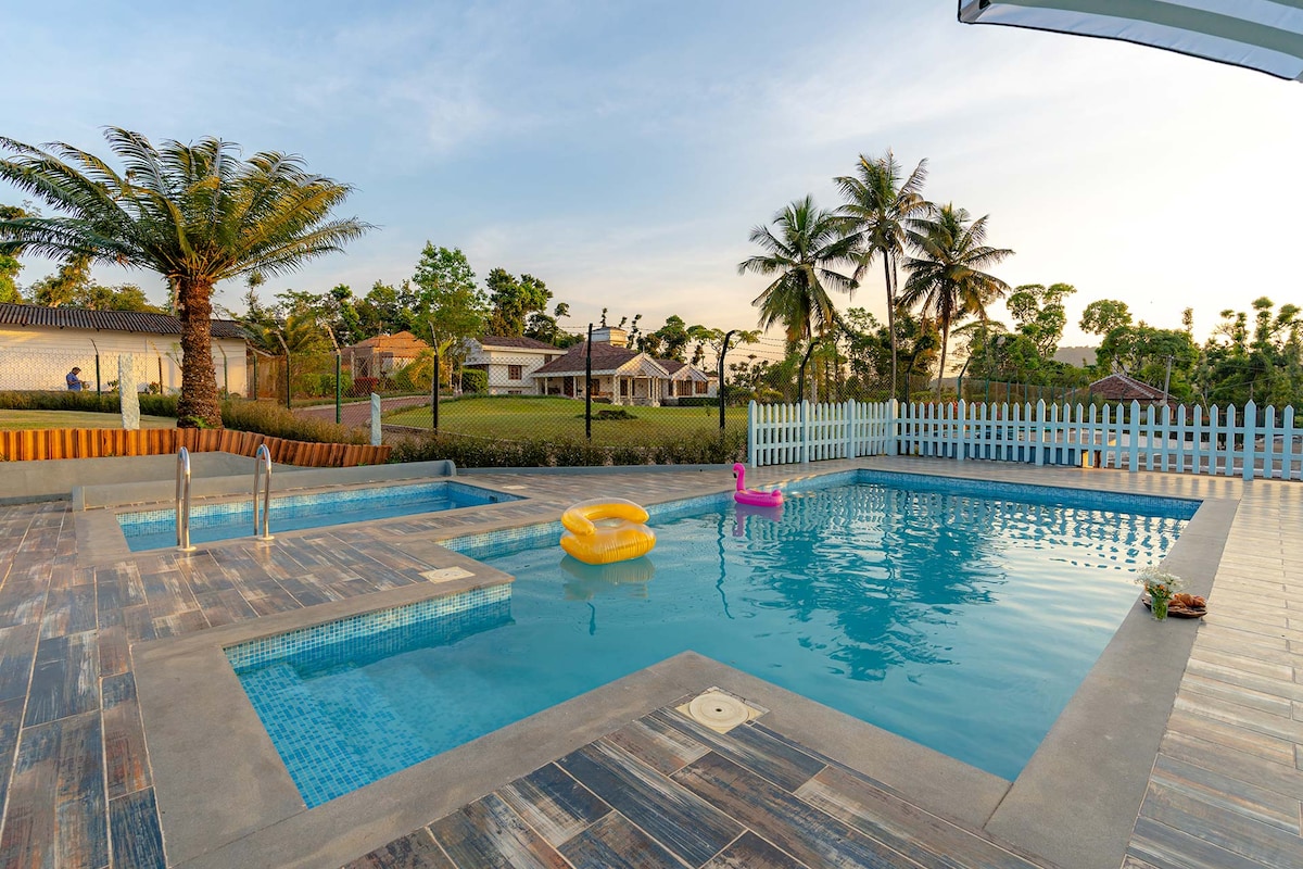 A spacious swimming pool area features two distinct pools, one designed for adults and a smaller one for children. Surrounding the pools are comfortable loungers and a clean deck area, with lush greenery and palm trees visible in the background.