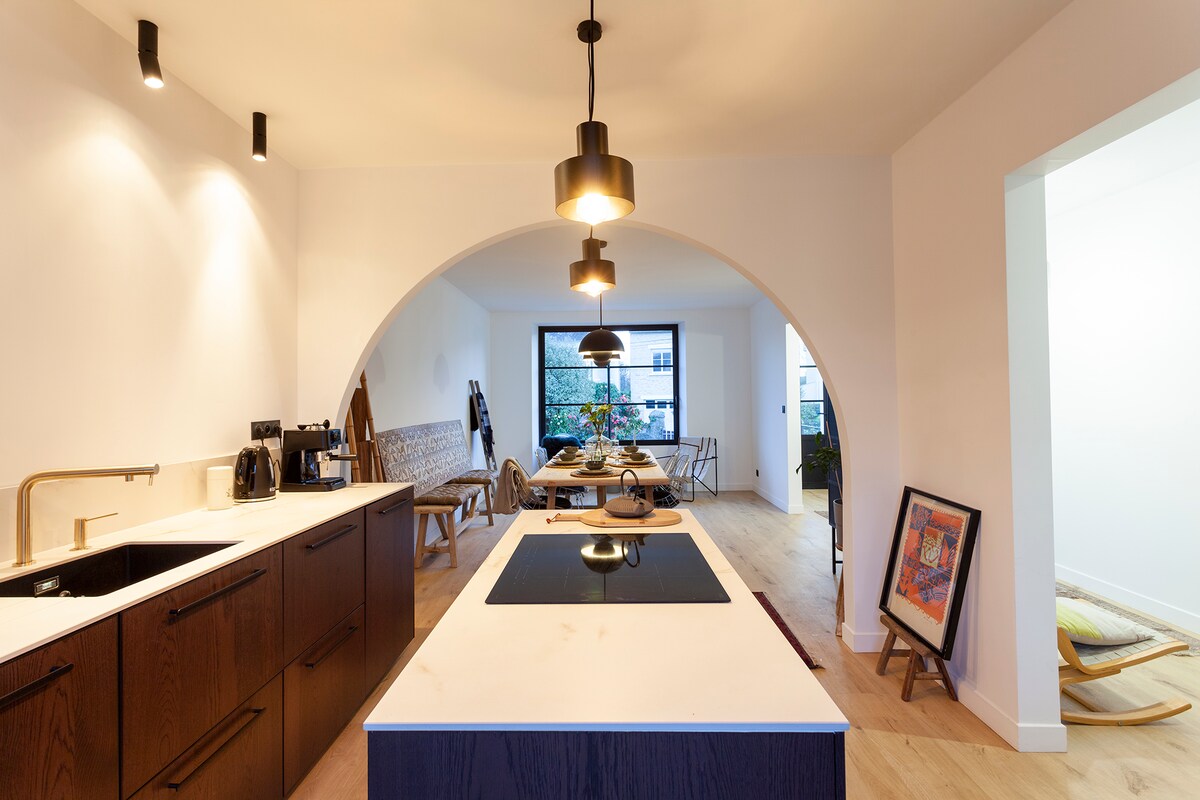 An open-plan kitchen area showcases a large island with a sleek cooktop, surrounded by dark wooden cabinetry. Elegant pendant lights hang from the ceiling, illuminating a spacious dining area in the background, defined by modern furniture and large windows allowing natural light.