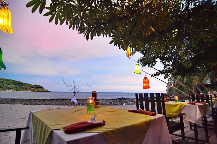 A beachfront restaurant is adorned with colorful lanterns hanging from trees, casting a soft glow over a wooden dining setup. Round tables with yellow tablecloths are set for dining, accompanied by candles. The ocean and beach can be seen in the background under a pastel sky.