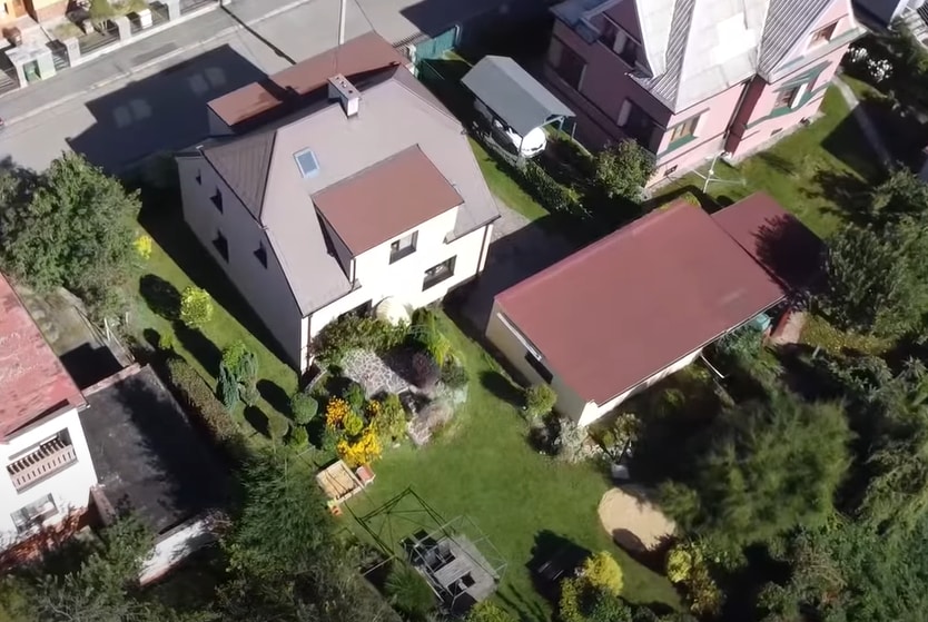 An aerial view showcases a family home surrounded by a well-maintained garden. A spacious lawn features a play area and flowerbeds, while parking is available beside the house. Nearby, other homes add to the neighborhood's charm.