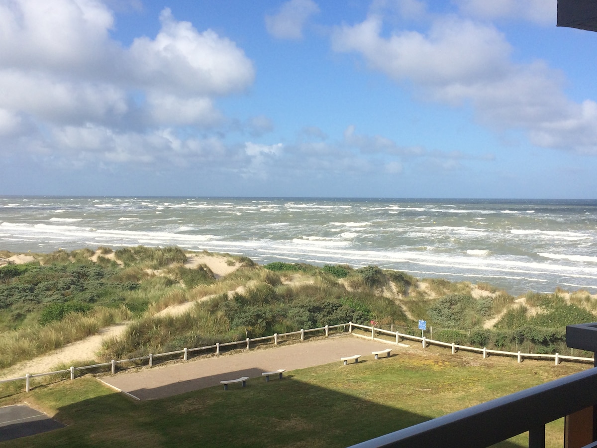 A panoramic view of the sea is displayed from a balcony, showcasing rolling waves meeting the sandy shore. The foreground features a grassy area bordered by a wooden fence, while the dunes are visible in the distance under a partly cloudy sky.