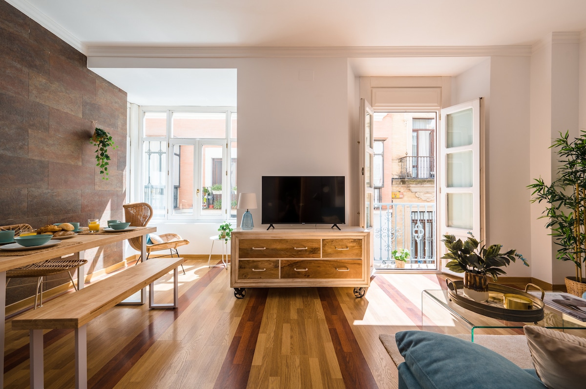 An inviting living area is showcased with wooden flooring and ample natural light from large windows. A low wooden table is set with breakfast items, while a modern TV sits on a stylish cabinet. Comfortable seating options include a blue sofa and wooden chairs.