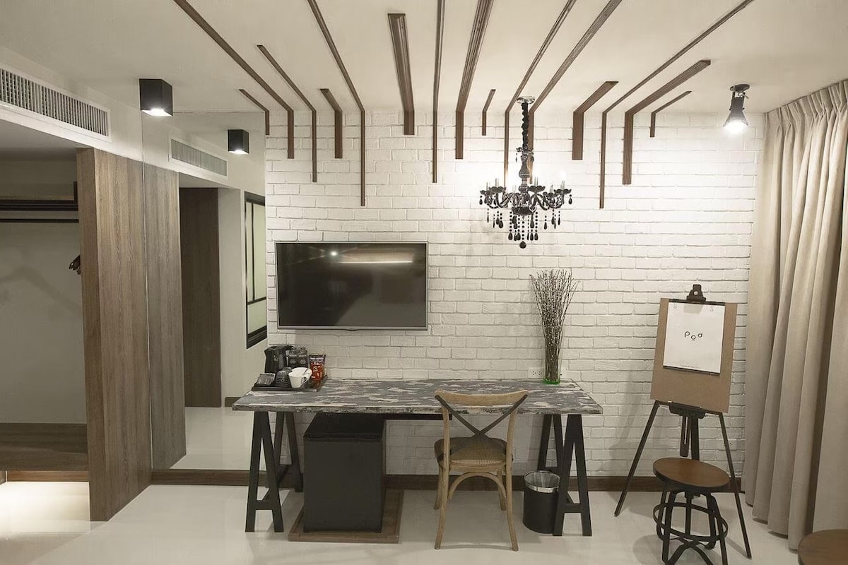 A stylish room features a wall-mounted flat screen TV and a contemporary desk with a chair. A decorative easel and a black stool are positioned nearby. Exposed wooden beams enhance the ceiling, and soft lighting adds a warm touch. A light-colored curtain partially drapes a window.