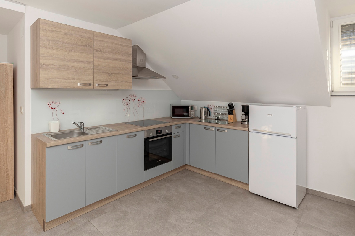 A modern kitchen displays sleek cabinetry in muted grey and natural wood tones. Equipped with essential appliances, including a refrigerator and microwave, the space offers a minimalist design with ample counter space. Soft lighting enhances the functionality of this welcoming cooking area.