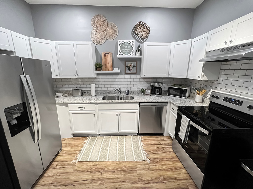 The kitchen features stainless steel appliances, including a refrigerator and oven, set against white cabinetry. A granite countertop provides ample workspace, while decorative items are displayed on the wall. A woven rug lies on the floor, adding texture to the space.