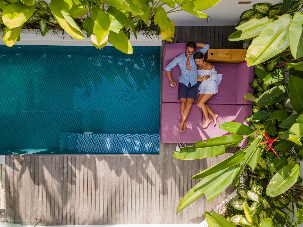 An aerial view reveals a private pool bordered by tropical greenery. A spacious purple lounging area accommodates two guests comfortably, with a wooden table alongside. Sunlight filters through the lush leaves, enhancing the peaceful ambiance of the outdoor space.