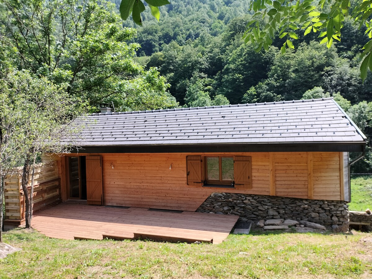 A charming wooden cabin is set against a backdrop of lush greenery. The exterior features a mix of stone and wood, with a sloped roof and shutters beside the windows. A spacious wooden deck extends in front, surrounded by grass and trees.