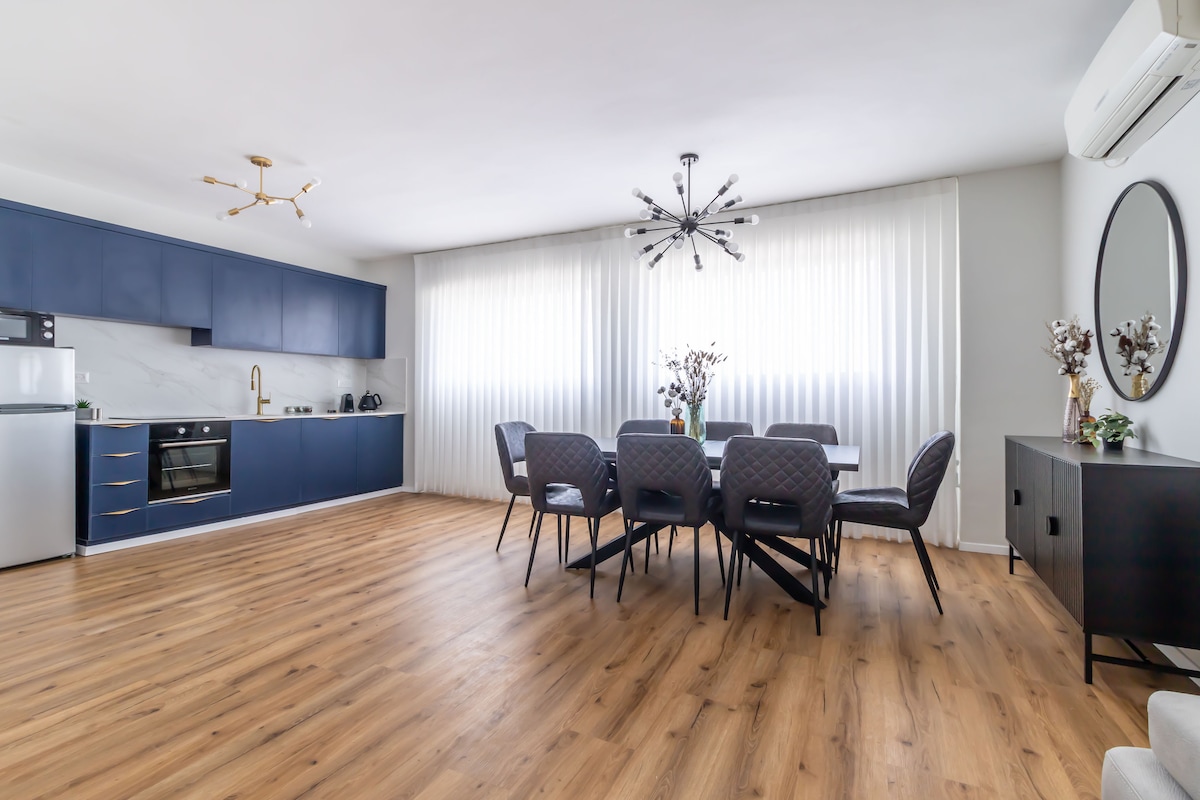 A modern dining area and kitchen are showcased, featuring a long dining table surrounded by stylish chairs. A sleek kitchen counter runs along one wall, and a range of appliances are visible. Natural light filters through large, sheer curtains, illuminating the warm wooden flooring.