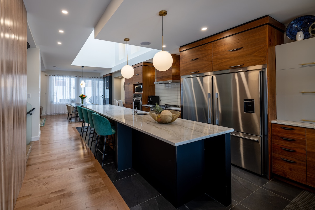 A spacious kitchen features a large island with a marble countertop and modern seating. Stainless steel appliances, including a refrigerator and oven, complement the wooden cabinetry. Soft lighting from pendant fixtures adds warmth, while natural light filters through large windows, enhancing the inviting ambiance.
