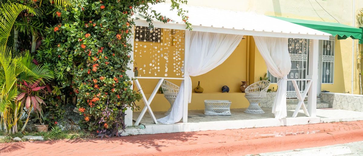 A shaded outdoor seating area is framed by lush greenery and colorful flowers. Two comfortable white wicker chairs are positioned under a white canopy, complemented by sheer curtains fluttering gently. The vibrant yellow wall of the nearby structure provides a bright backdrop.